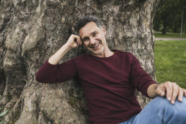 Happy mature man leaning on tree trunk in park - UUF26242