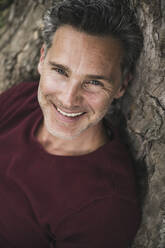 Smiling mature man leaning on tree trunk - UUF26244