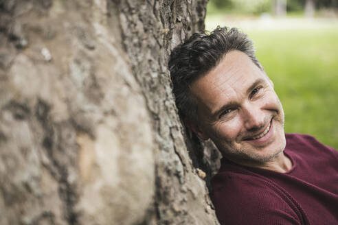Lächelnder reifer Mann lehnt sich im Park an einen Baumstamm - UUF26248