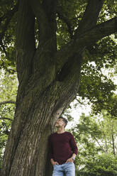 Glücklicher Mann mit Händen in den Taschen an einem Baum im Park stehend - UUF26254