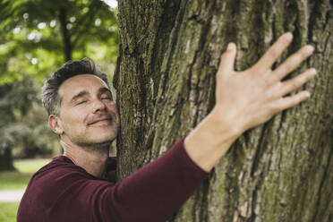 Lächelnder Mann mit geschlossenen Augen, der einen Baum im Park umarmt - UUF26258