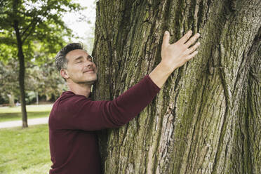 Lächelnder reifer Mann mit geschlossenen Augen, der einen Baum im Park umarmt - UUF26259