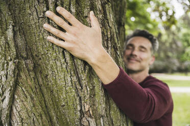 Happy mature man with eyes closed hugging tree at park - UUF26260