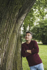 Mature man with arms crossed leaning on tree trunk - UUF26263