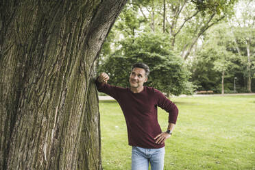 Smiling mature man with hand on hip looking at tree in park - UUF26267