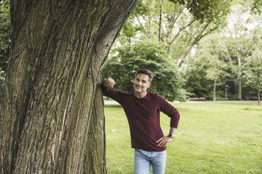 Glücklicher reifer Mann mit Hand auf der Hüfte stehend an einem Baum im Park - UUF26268