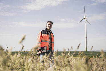 Engineer walking at field near wind turbine on sunny day - UUF26308