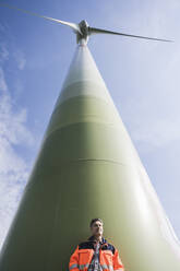 Engineer standing in front of tall wind turbine on sunny day - UUF26312