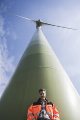 Smiling engineer standing in front of tall wind turbine on sunny day - UUF26314