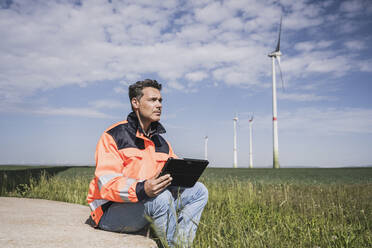 Engineer with tablet PC sitting near wind turbine on field - UUF26315