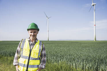 Lächelnder Ingenieur mit Schutzhelm vor einer Windkraftanlage - UUF26327