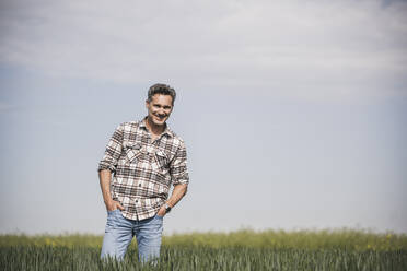 Glücklicher reifer Mann mit Händen in den Taschen auf einem Feld an einem sonnigen Tag - UUF26331