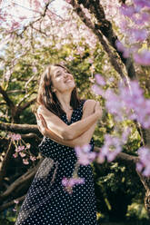 Lächelnde junge Frau mit geschlossenen Augen, die sich im Garten umarmt - OMIF00851