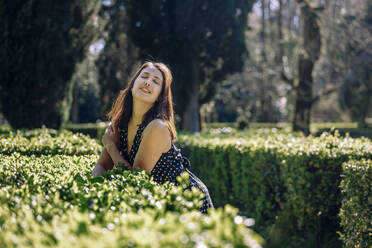 Frau mit geschlossenen Augen lehnt an einer Hecke im Garten an einem sonnigen Tag - OMIF00865