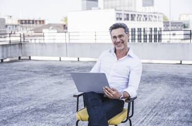 Lächelnder Geschäftsmann mit Laptop auf der Terrasse eines Gebäudes sitzend - UUF26350