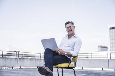 Älterer Geschäftsmann mit Brille sitzt mit Laptop auf einem Stuhl - UUF26352