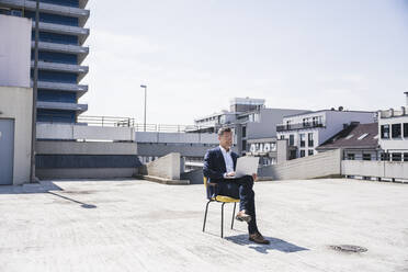 Smiling businessman with laptop sitting on chair - UUF26365