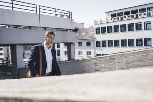 Geschäftsmann mit In-Ear-Kopfhörern beim Spaziergang in einem Parkhaus an einem sonnigen Tag - UUF26374