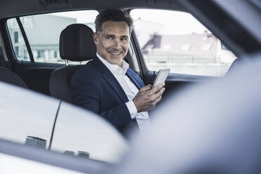 Smiling businessman sitting with smart phone in car - UUF26394