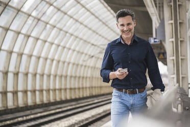 Lächelnder reifer Mann mit Smartphone beim Spaziergang am Bahnhof - UUF26402