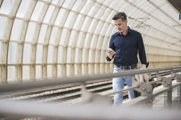 Älterer Mann, der ein Smartphone benutzt und am Bahnhof spazieren geht - UUF26407