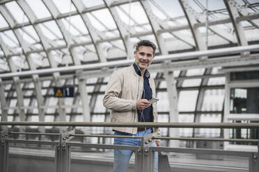 Lächelnder Geschäftsmann mit Smartphone und Tasche beim Spaziergang am Bahnhof - UUF26422