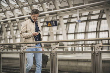 Älterer Geschäftsmann, der ein Smartphone benutzt und mit einer Tasche am Bahnhof spazieren geht - UUF26423