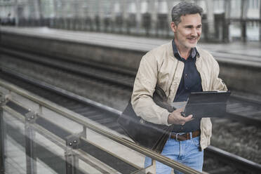 Lächelnder Geschäftsmann mit Tablet-PC und Tasche auf dem Bahnhof - UUF26425