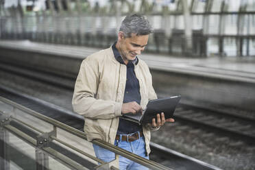 Smiling businessman using tablet PC standing at railroad station - UUF26426