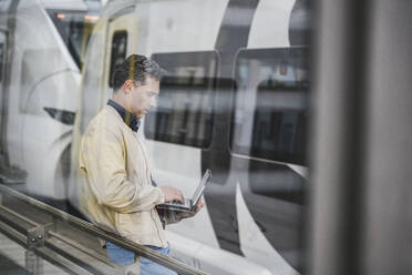 Businessman using tablet PC standing in front of train at railroad station - UUF26427