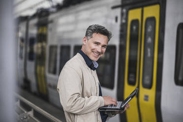 Happy businessman with tablet PC standing in front of train at railroad station - UUF26428
