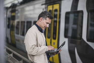 Mature businessman using tablet PC in front of train at railroad station - UUF26429