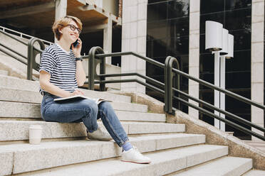 Lächelnde Geschäftsfrau, die auf einer Treppe sitzend mit einem Smartphone spricht - MRRF02204