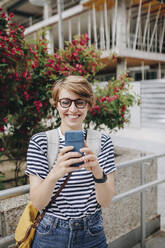 Smiling woman using smart phone standing in front of plants - MRRF02213