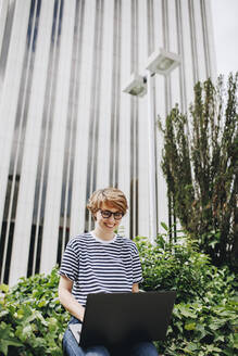 Woman using laptop sitting in front of building - MRRF02216