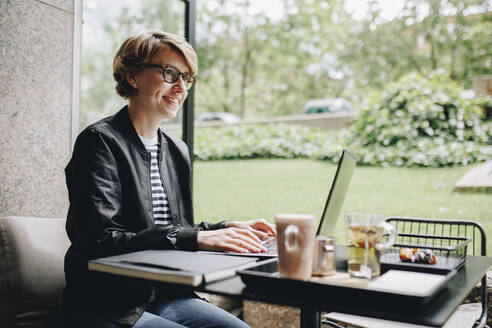 Lächelnde Geschäftsfrau mit Laptop in einem Café sitzend - MRRF02240