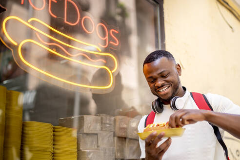 Glücklicher junger Mann isst Hot Dog und lehnt sich an ein Restaurantfenster - OCMF02415