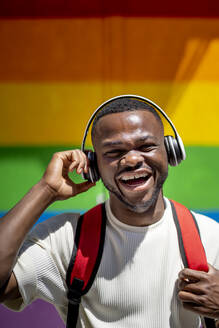 Glücklicher Mann mit drahtlosen Kopfhörern vor einer Regenbogenwand - OCMF02419