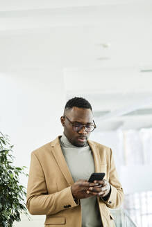 Young businessman wearing eyeglasses surfing net through mobile phone in office - DSHF00442