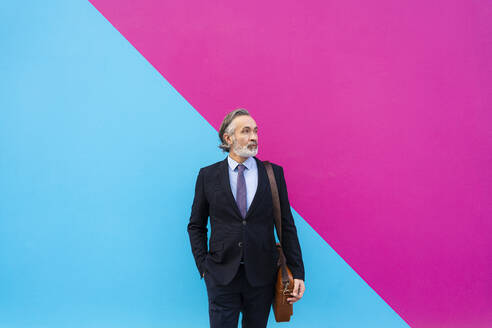 Älterer Geschäftsmann mit Tasche vor rosa und blauer Wand stehend - OIPF02007