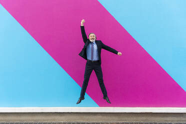 Glücklicher reifer Geschäftsmann mit erhobener Hand, der vor einer rosa und blauen Wand springt - OIPF02022