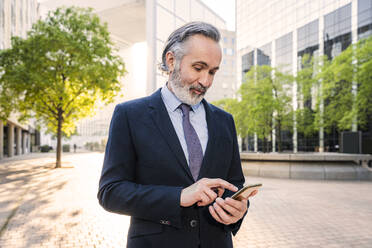 Älterer Geschäftsmann mit Smartphone im Büropark stehend - OIPF02054