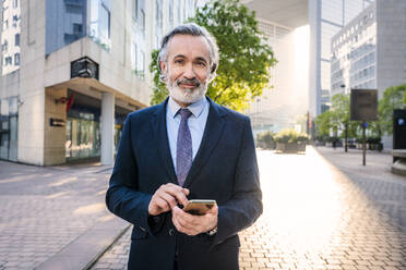 Lächelnder Geschäftsmann mit Smartphone im Büropark stehend - OIPF02056
