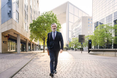 Älterer Geschäftsmann mit Tasche zu Fuß im Büropark an einem sonnigen Tag - OIPF02057