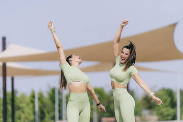 Cheerful twin sisters with hands raised enjoying sunny day - OCMF02429