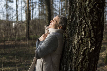 Woman with closed eyes leaning against tree trunk - LLUF00591