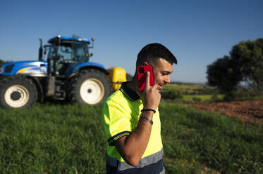Lächelnder Bauer, der vor einem Traktor mit seinem Handy telefoniert - JCMF02258