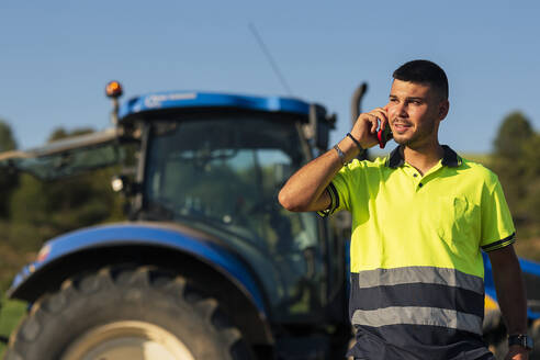 Junger Landwirt spricht mit seinem Smartphone vor einem Traktor - JCMF02268