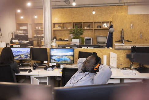 Geschäftsmann mit Headset, der sich mit den Händen hinter dem Kopf im Büro zurücklehnt - CAIF32846