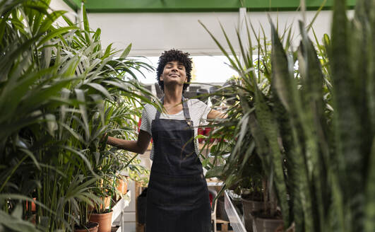 Smiling owner with eyes closed standing amidst plants at nursery - JCCMF06469